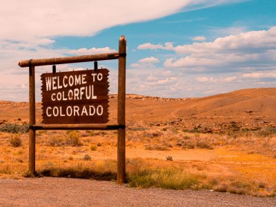 "Welcome to Colorado" sign on the left of the screen in front of a large expanse of yellow grass