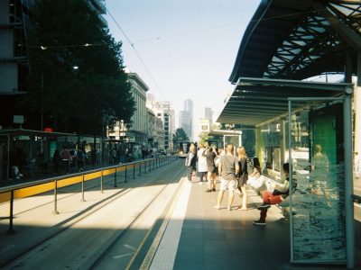 Image of people waiting at an urban train stop. Large buildings can be seen in the background.